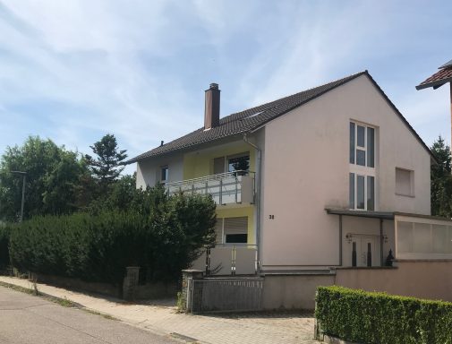 Helles Haus mit Balkon, umgeben von Bäumen und einer niedrigen Hecke.